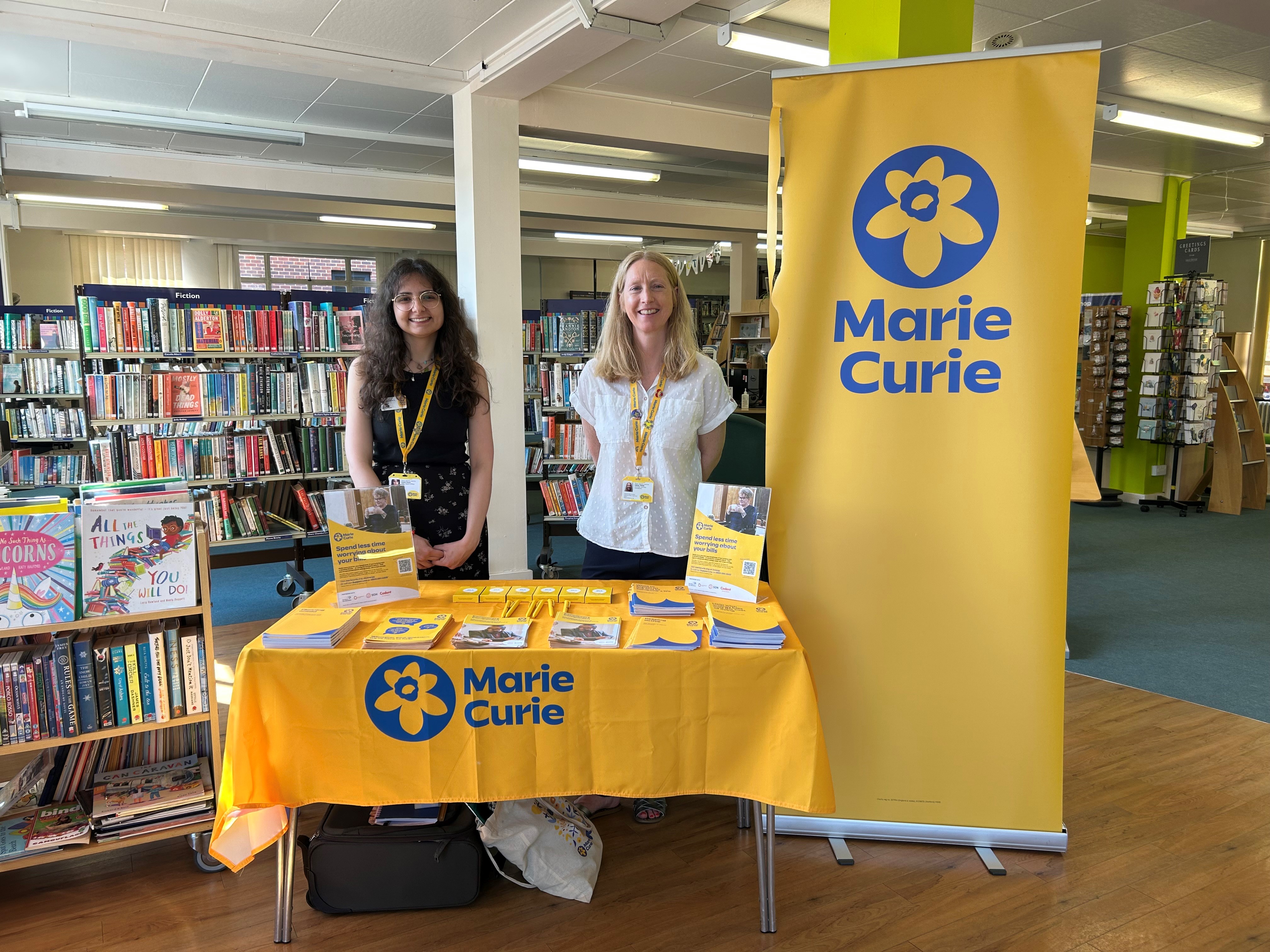 Two women standing behind a promotional table display for Marie Curie in a library.