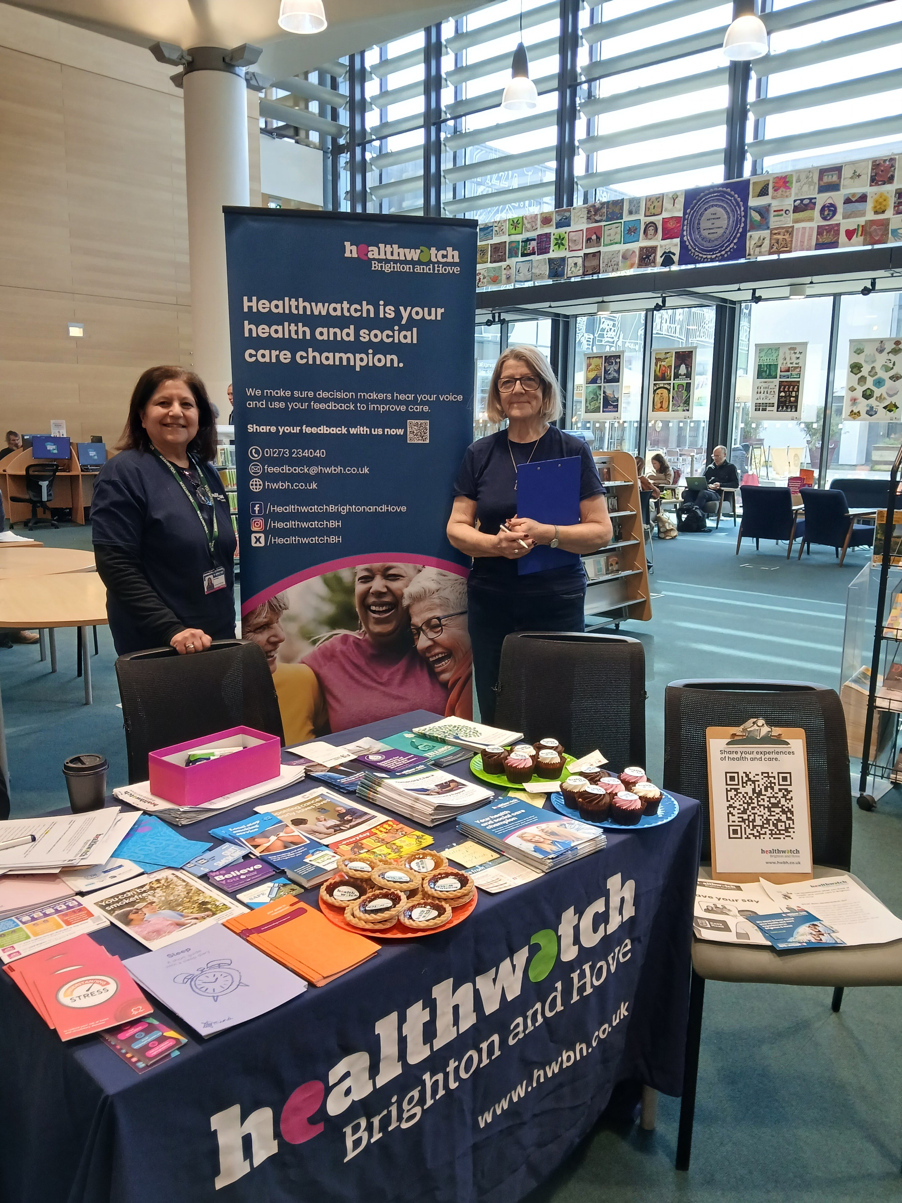 Two volunteers stood in front of a Healthwatch Brighton and Hove stall in an indoor setting.
