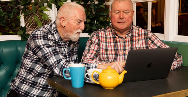 Two older men helping each other to use a laptop while sat in a cafe.