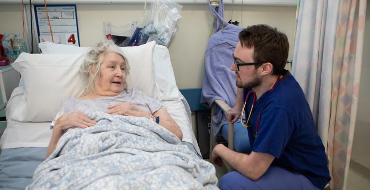 Older woman in hospital bed receiving care from a male doctor.