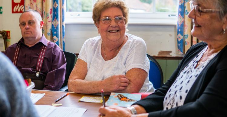 Three older adults sat around a table together talking and smiling.