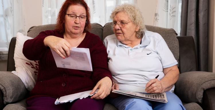 Two older women sat on a sofa looking at an official letter while one takes notes with a pen.