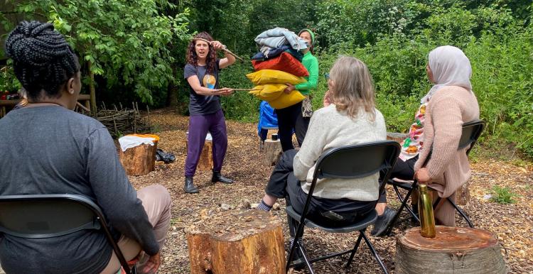 Three people sat on chairs and two standing in an outdoor, wooded setting
