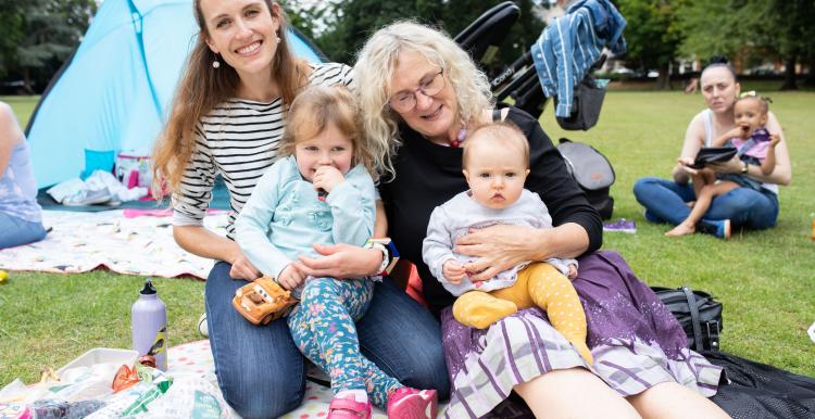 Two women with a small child and baby sat on a picnic blanket outdoors.