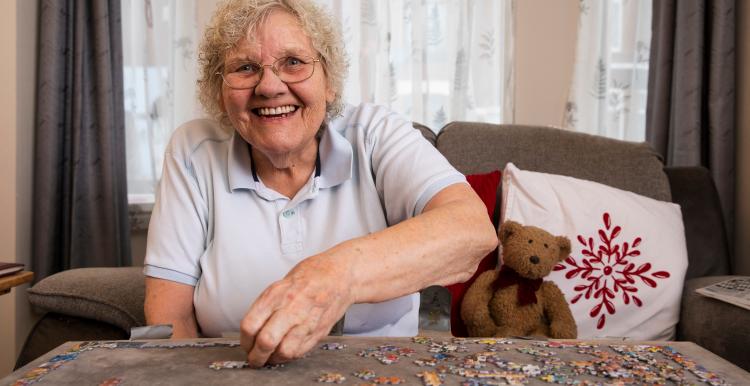 Older woman smiling while doing a puzzle in a living room.