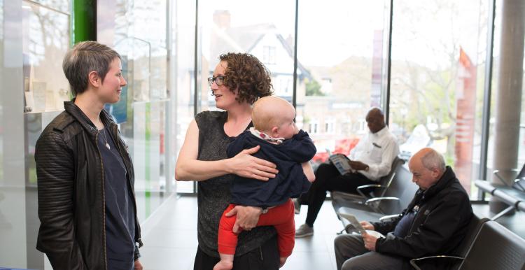 Two women talking while holding a baby in a clinical waiting room