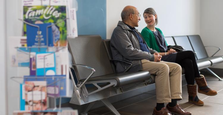 Two people talking while sat down in a GP surgery waiting area.