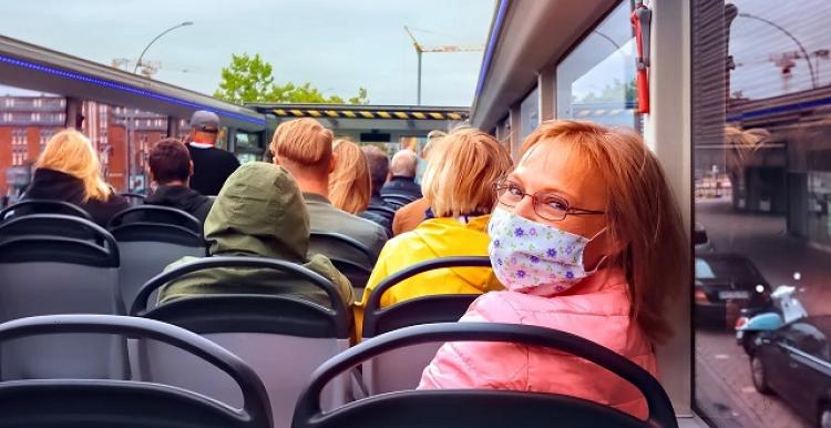 Woman on top deack of open top bus, looking back at us, wearing face mask.