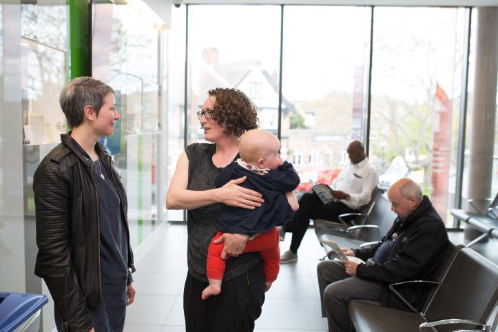 Two women talking while holding a baby in a clinical waiting room