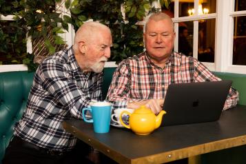 Two older men helping each other to use a laptop while sat in a cafe.