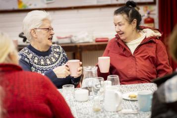 Two older women talking sat down at a communal table drinking hot drinks and talking.