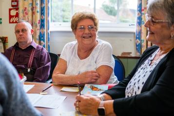 Three older adults sat around a table together talking and smiling.