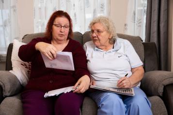 Two older women sat on a sofa looking at an official letter while one takes notes with a pen.