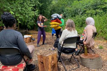 Three people sat on chairs and two standing in an outdoor, wooded setting