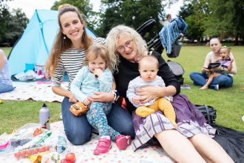 Two women with a small child and baby sat on a picnic blanket outdoors.
