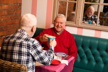 Two men sat in a café smiling with hot drinks