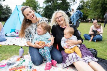 Two women holding small children while sat outside in a park setting.