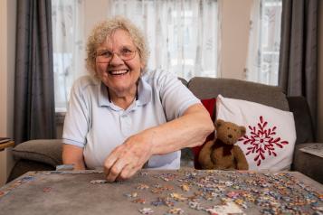 Older woman smiling while doing a puzzle in a living room.