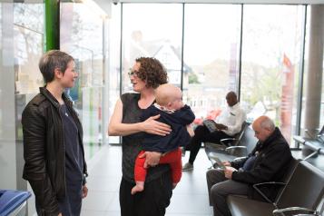 Two women talking while holding a baby in a clinical waiting room