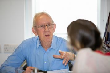 older man facing us across a desk speaking to a woman with her back to us