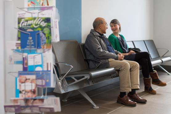 Two people talking while sat down in a GP surgery waiting area.