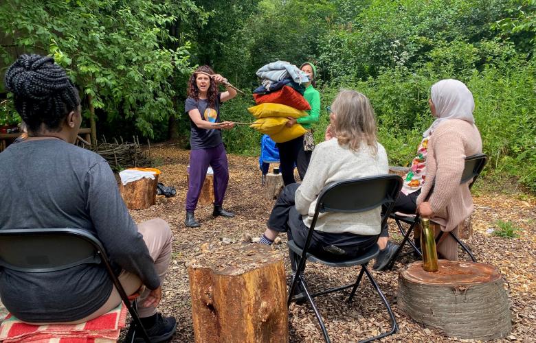 Three people sat on chairs and two standing in an outdoor, wooded setting