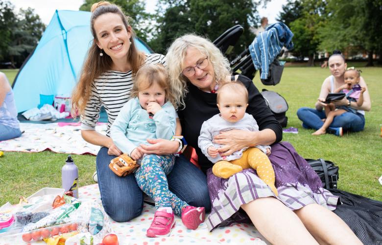 Two women with a small child and baby sat on a picnic blanket outdoors.