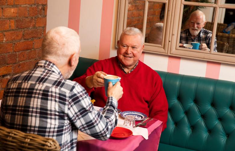 Two men sat in a café smiling with hot drinks