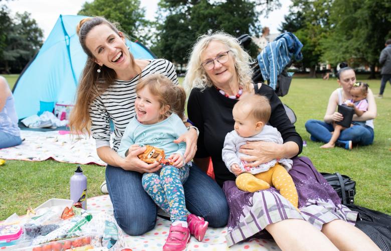 Two women holding small children while sat outside in a park setting.