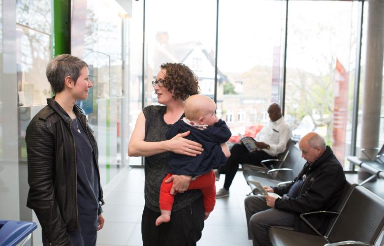 Two women talking while holding a baby in a clinical waiting room