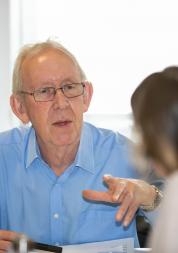 older man facing us across a desk speaking to a woman with her back to us